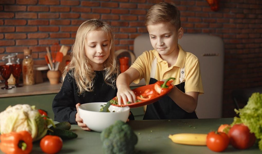 Little girl and boy mixing cutted pepper with other fresh ingredients for salad in big bowl while cooking at table with various fresh vegetables and fruits