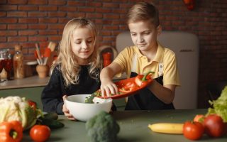 Little girl and boy mixing cutted pepper with other fresh ingredients for salad in big bowl while cooking at table with various fresh vegetables and fruits