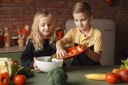 Little girl and boy mixing cutted pepper with other fresh ingredients for salad in big bowl while cooking at table with various fresh vegetables and fruits